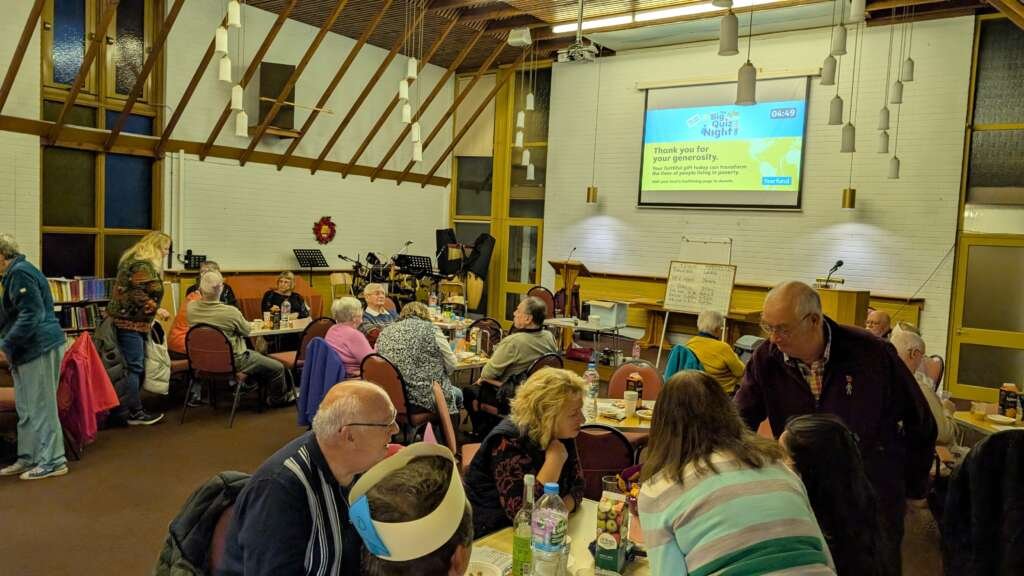 Community hall scene with seniors seated at round tables for a social event; projector screen shows 'Big Quiz Night' and a wreath decorates the wall in the background.