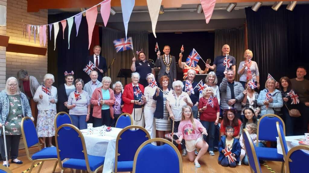 Group of people of mixed ages posing for a photo indoors at a patriotic event, many waving Union Jack flags under colorful bunting on a stage.