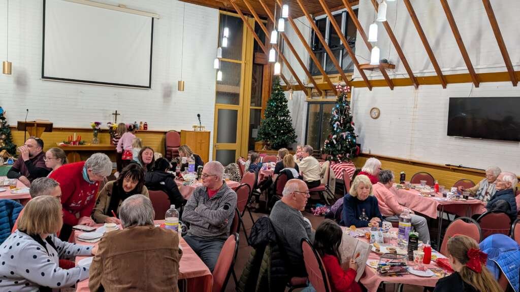 Community Christmas party in a church hall with many people seated at round tables, eating and chatting near Christmas trees and decorations