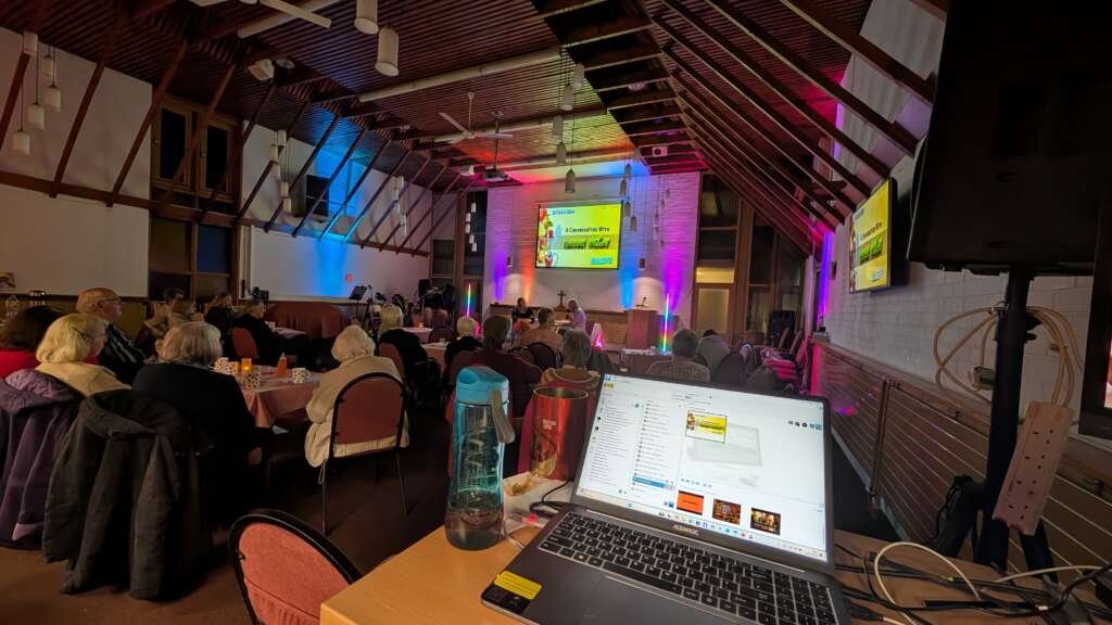 Audience seated at round tables in a church hall watching a presentation on a stage with colorful lighting.