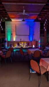 Stage area of a church or event space with a cross on the altar, two chairs, a projector screen, and colorful uplighting creating a vibrant atmosphere.