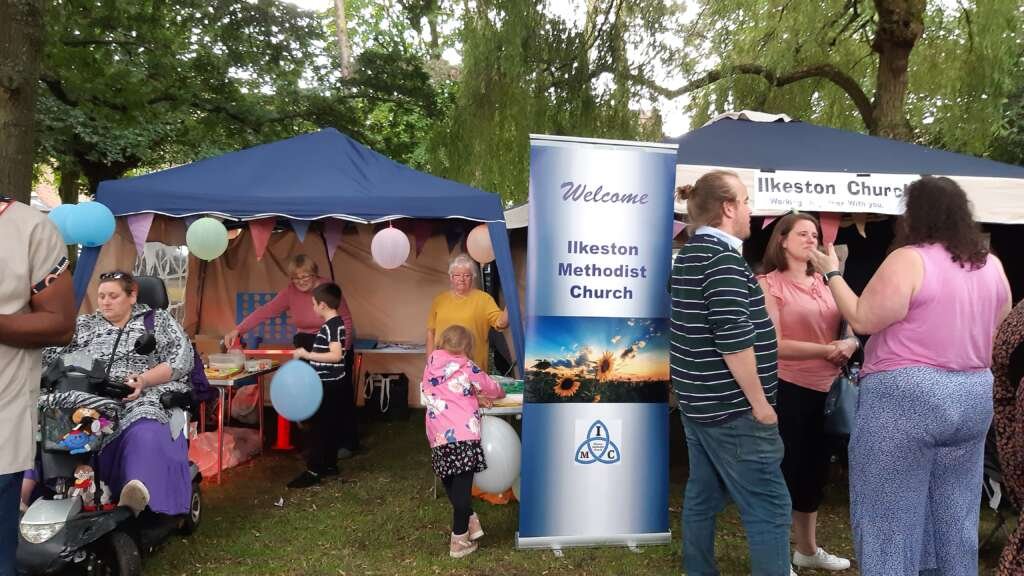 Outdoor community fair with blue tents, bunting, and balloons near Ilkeston Methodist Church banner.
